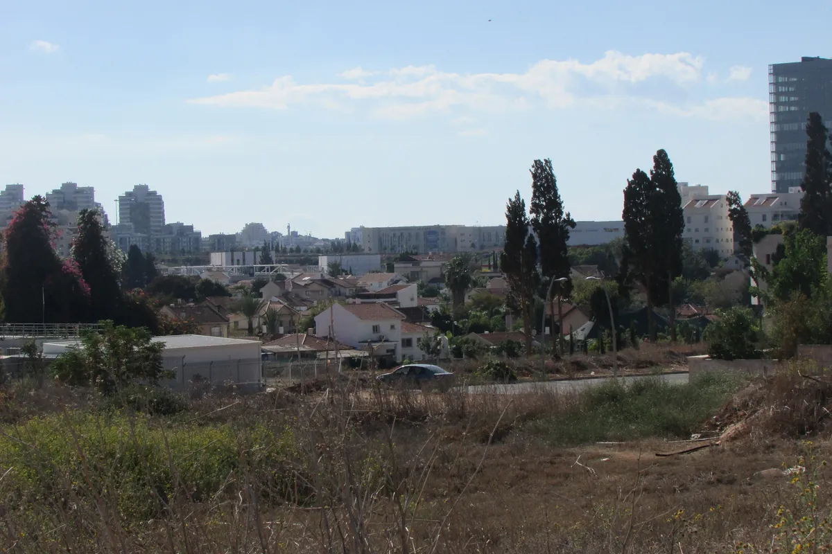 Open ground and low houses in the Aliya neighbourhood of Kfar Saba, with tower blocks behind